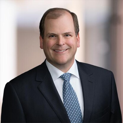 A man in a dark pinstripe suit, white shirt, and patterned tie is smiling at the camera against a blurred indoor background of a corporate law office.