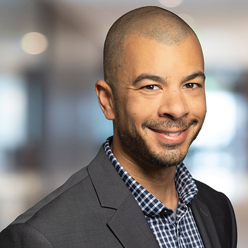 A man with a shaved head, trimmed beard, and a checked shirt under a dark blazer smiles at the camera against a blurred indoor background, reflecting the professionalism found in top law offices and among lawyers in Chicago.