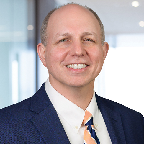 Smiling man in a blue suit jacket, white shirt, and striped tie, standing in an office setting with blurred background—ideal for promoting law offices or litigation support services.