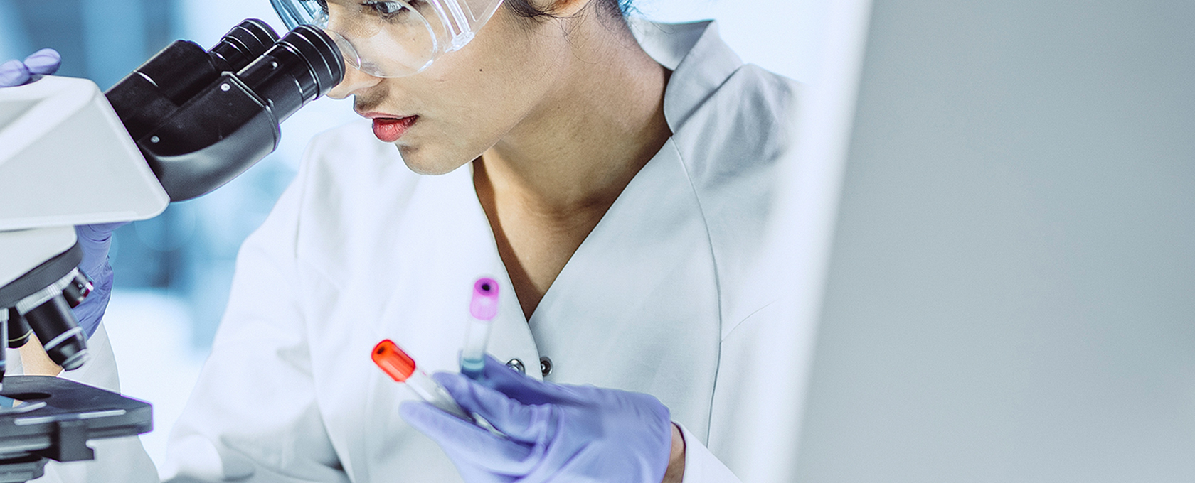 A scientist wearing protective glasses examines samples under a microscope while holding two test tubes in a laboratory, providing crucial research that can aid intellectual property law and litigation support for lawyers in Chicago.