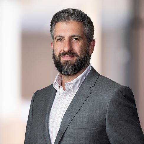 A man with gray hair and a beard, dressed in a gray suit jacket and white shirt, stands against a blurred indoor background—evoking the professional atmosphere of law offices or a corporate law office.