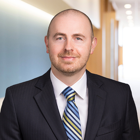A man in a dark suit, striped tie, and white shirt stands in a brightly lit corporate law office hallway, looking at the camera with a slight smile.