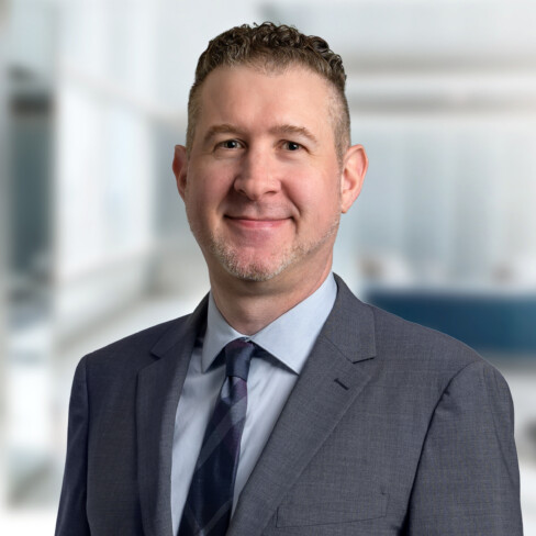 A man in a gray suit and tie smiles at the camera, standing in a modern office setting with blurred background—reflecting the professionalism of Chicago lawyers experienced in intellectual property law.