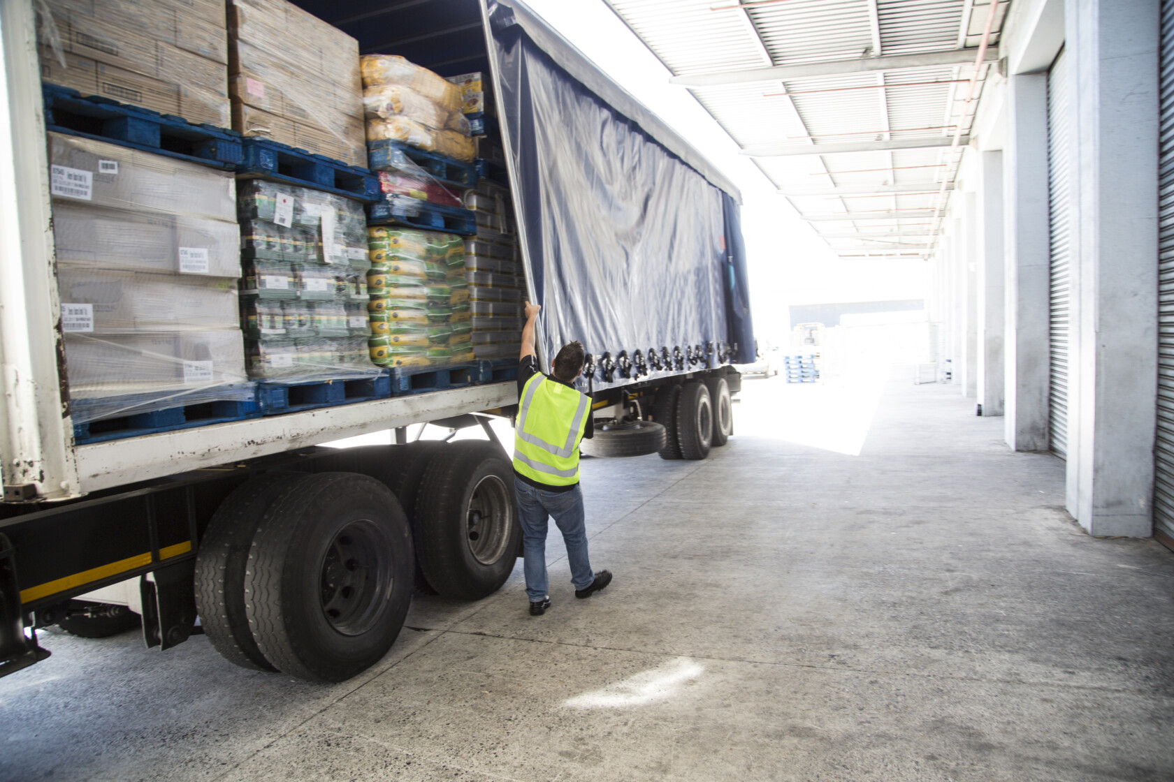 A worker in a high-visibility vest opens the side curtain of a loaded delivery truck at a warehouse loading dock.