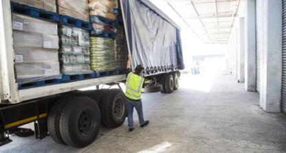 A worker in a high-visibility vest opens the side curtain of a loaded delivery truck at a warehouse loading dock.