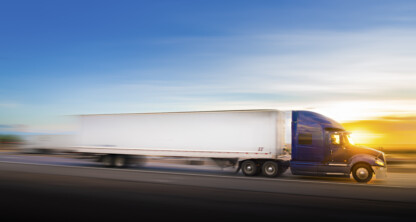 A blue semi-truck with a white trailer drives quickly on a highway at sunset, with a blurred background indicating motion.