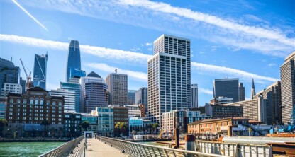 View of downtown San Francisco skyline from a pier, with tall modern skyscrapers, blue sky, and wispy clouds overhead.