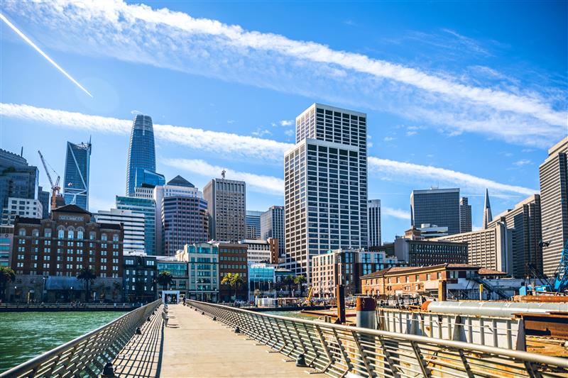 View of downtown San Francisco skyline from a pier, with tall modern skyscrapers, blue sky, and wispy clouds overhead.
