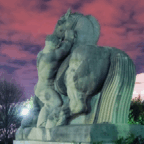 Stone statue of a person and a horse under a purple and pink cloudy sky, with trees and a building visible in the background.