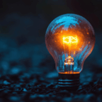 A lit incandescent light bulb sits on a dark surface surrounded by small pebbles, glowing with a warm orange light against a blue-tinted background.