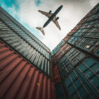 A commercial airplane flies overhead between tall stacks of colorful shipping containers under a cloudy sky.