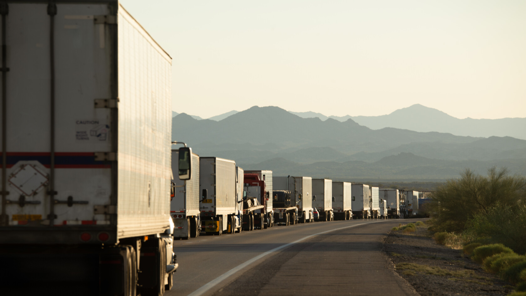 A long line of semi-trailer trucks is stopped along a highway with mountains visible in the background under a clear sky.