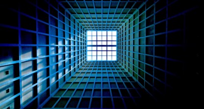A geometric grid structure viewed from below, with blue and green tones, leading towards a bright white square opening at the top center.