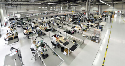 Large, brightly lit factory floor with multiple workstations where people assemble electronic components at long tables, surrounded by industrial equipment and supplies.