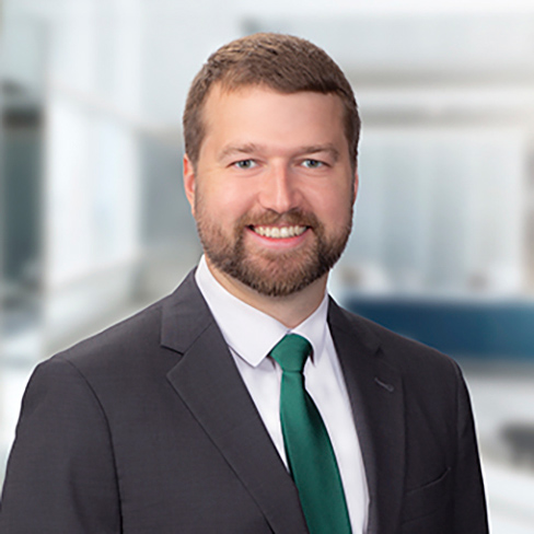 A man with short brown hair and a beard, wearing a dark suit, white shirt, and green tie, smiles at the camera in a modern office setting.