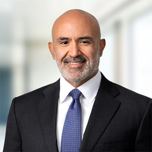 A bald man with a beard, wearing a dark suit, white shirt, and blue tie, stands in front of a blurred office background.