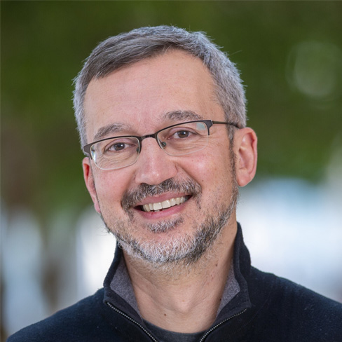 A middle-aged man with short gray hair, glasses, and a trimmed beard is smiling, wearing a dark zip-up sweater, with a blurred green background.