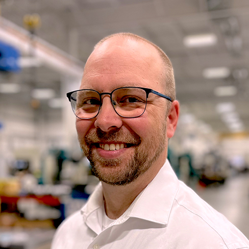A man with glasses, a beard, and a white shirt smiling in an indoor industrial or laboratory setting.