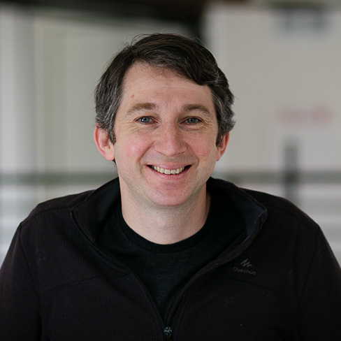 A man with short brown hair and blue eyes is smiling at the camera, wearing a dark zip-up jacket over a black shirt, with a blurred indoor background.