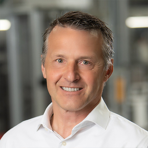 A middle-aged man in a white collared shirt smiles at the camera with a blurred indoor background.