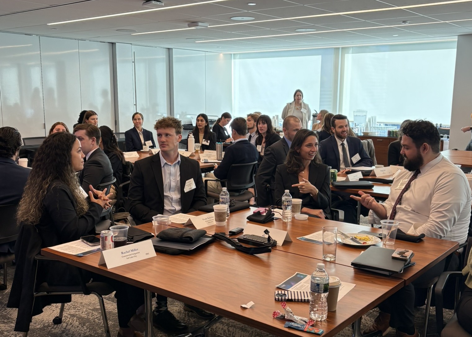 A group of professionals in business attire engage in conversation around tables in a modern conference room with large windows.