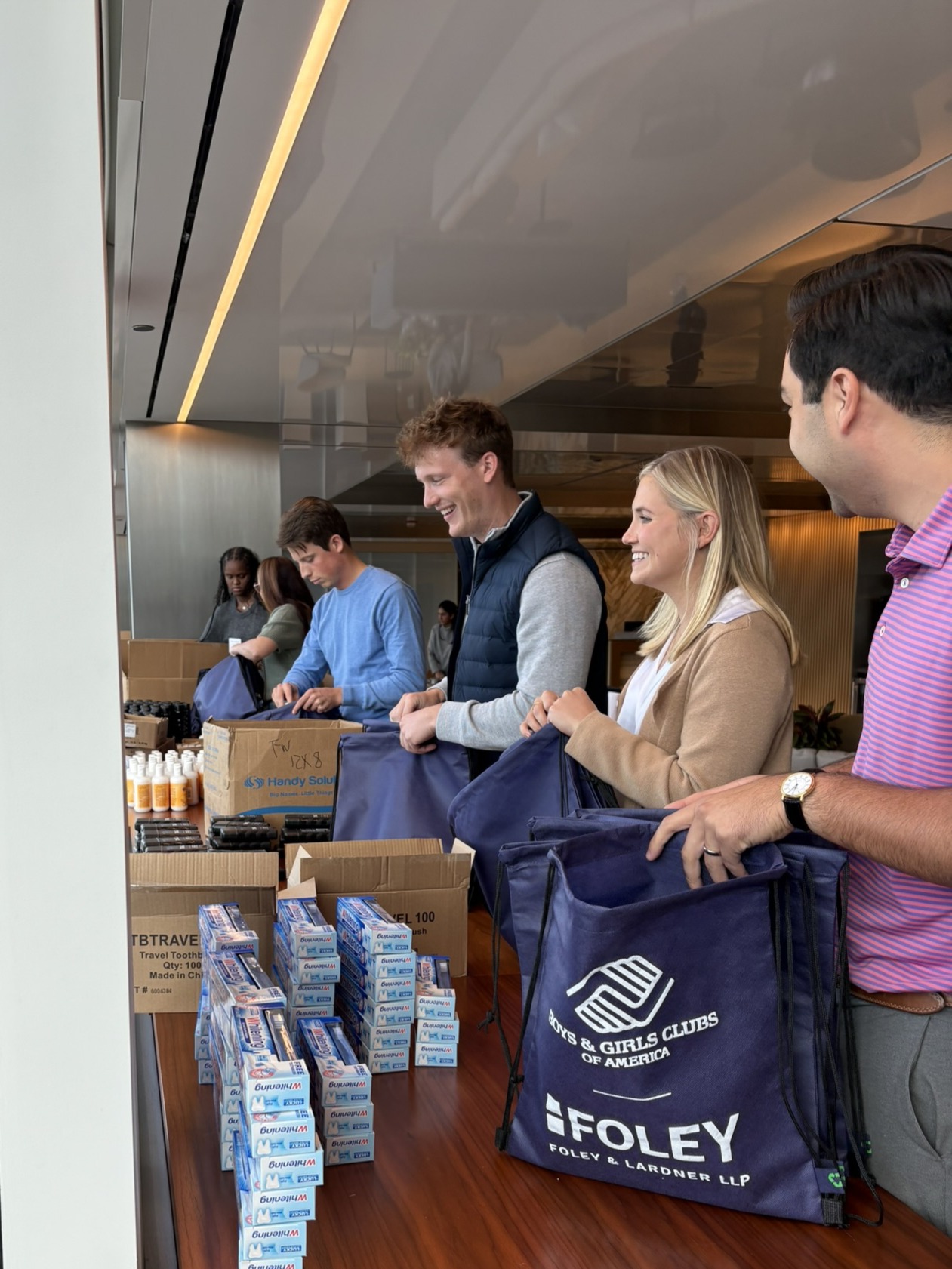 Four people stand at a table packing toiletries and hygiene products into Boys & Girls Clubs of America bags, with boxes of supplies visible in the foreground.