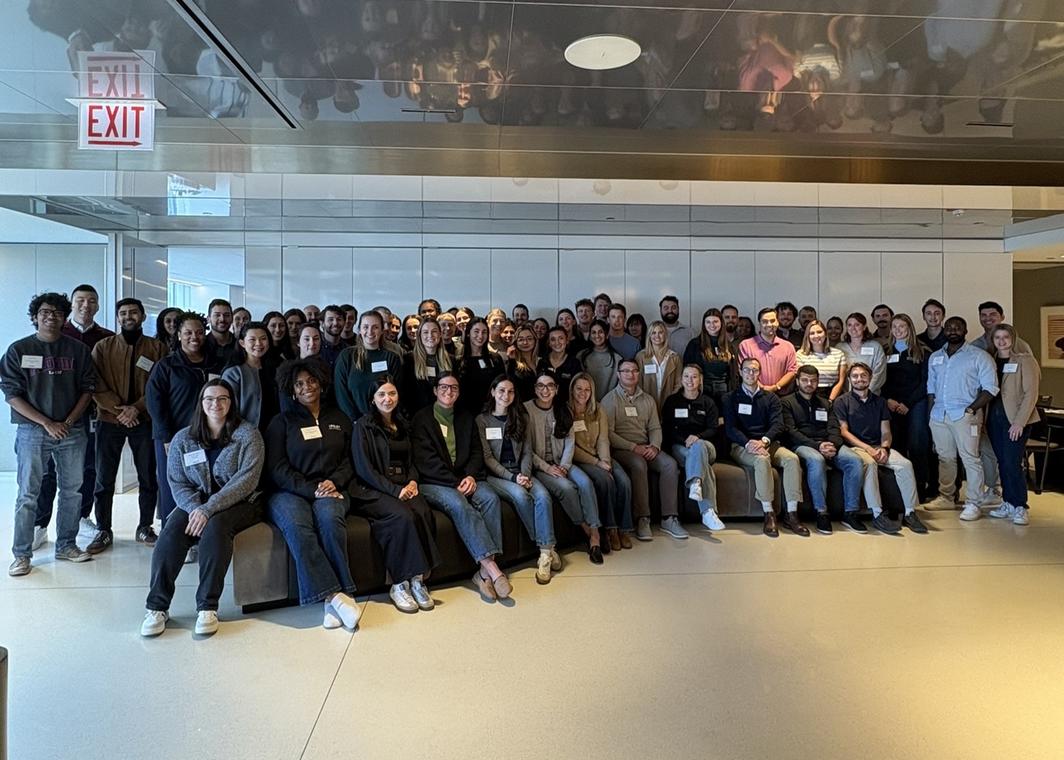 A large group of people, most wearing name tags, pose for a photo indoors in front of a reflective wall with exit signs overhead.