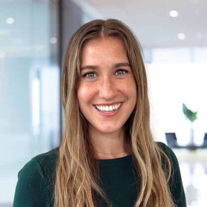 A woman with long, light brown hair wearing a dark green top is smiling in a modern, bright office setting.