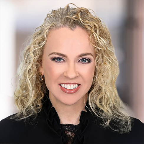 A woman with curly blonde hair, light skin, and blue eyes is smiling at the camera, wearing a black top. The background is softly blurred.