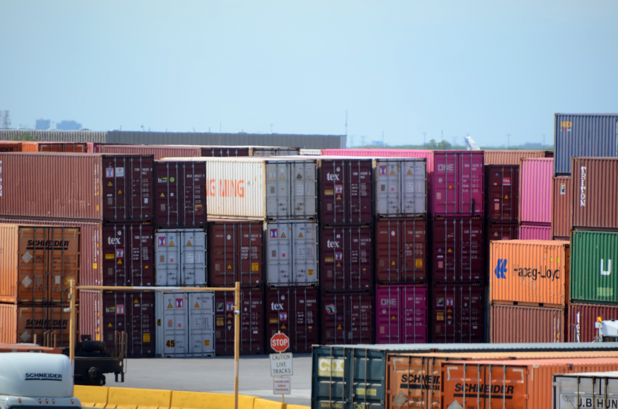 Stacked shipping containers in various colors are organized at a shipping yard, with a stop sign and traffic signs visible in the foreground.
