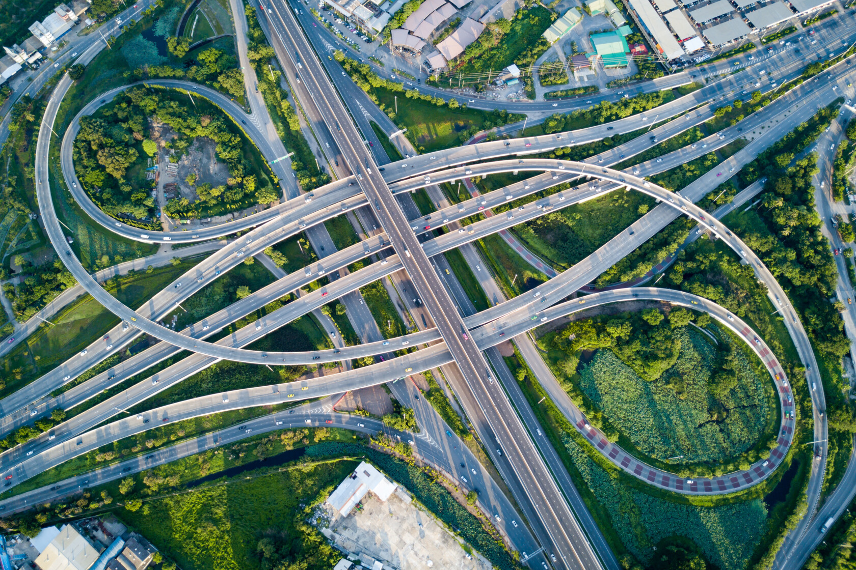 Aerial view of a complex highway interchange with multiple lanes, overpasses, and looping ramps surrounded by greenery and nearby buildings.