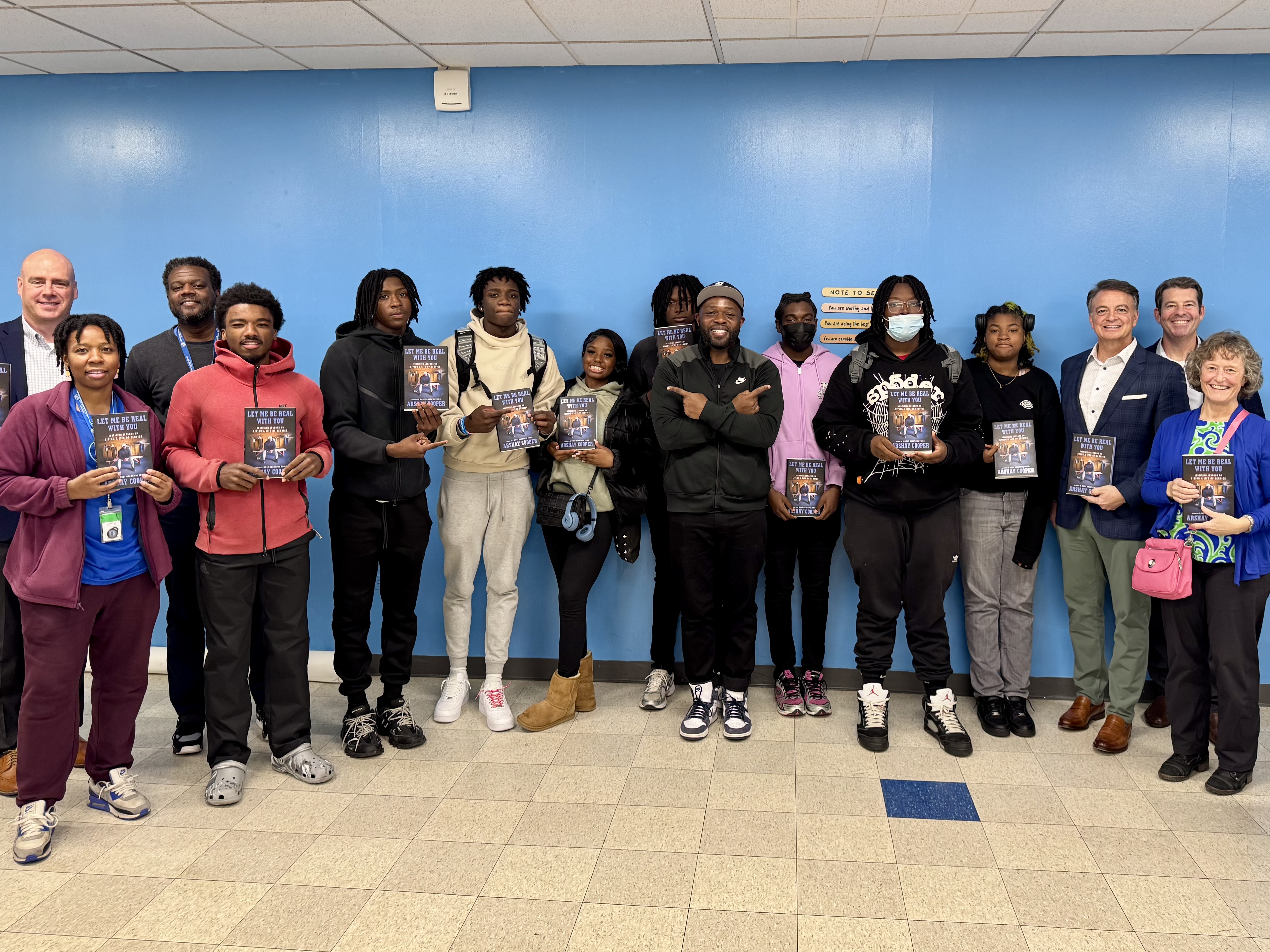 A group of people stand indoors against a blue wall, holding up copies of a book and looking at the camera.