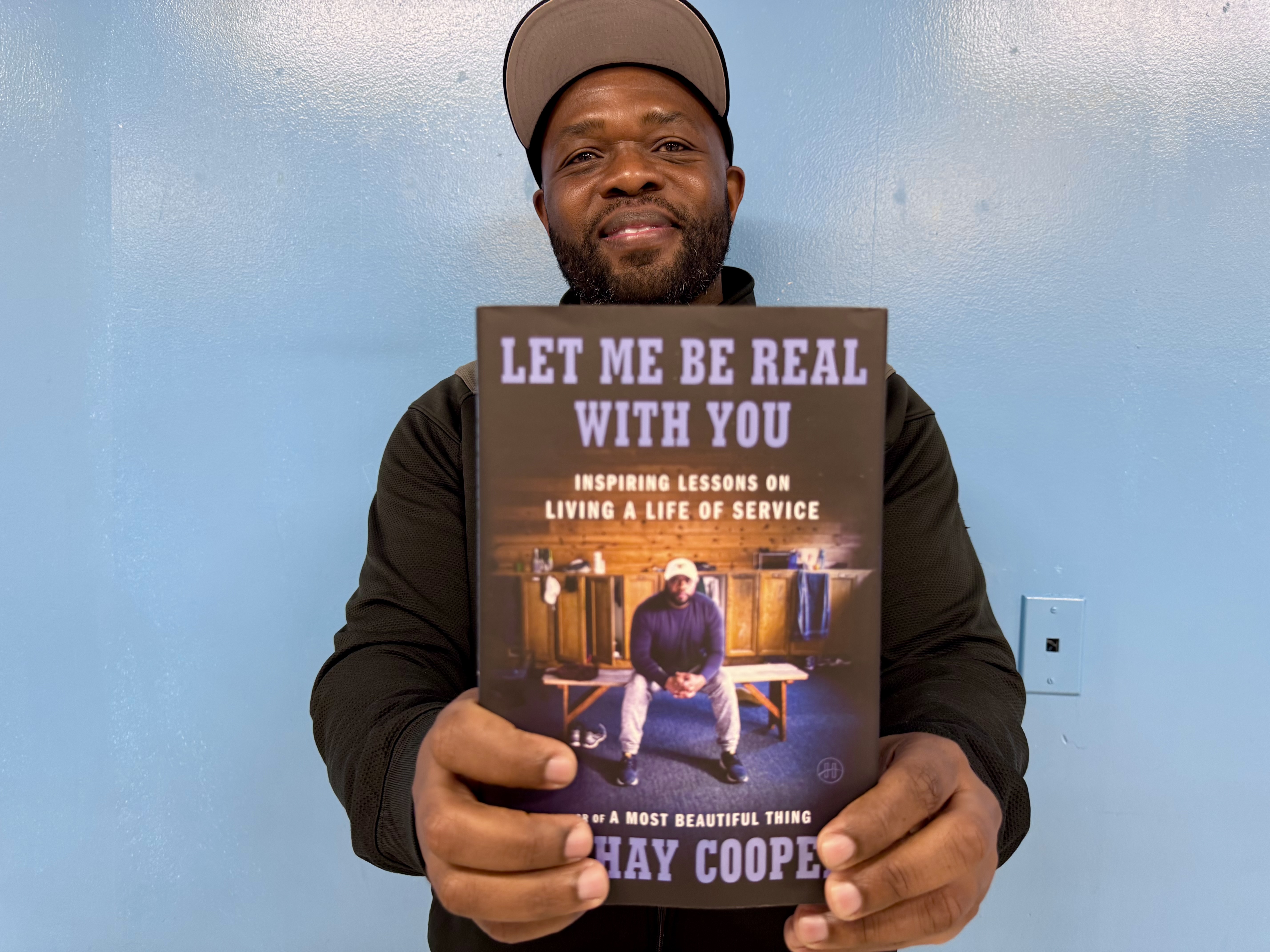 A man in a dark jacket and cap holds up a book titled "Let Me Be Real With You" against a light blue wall.