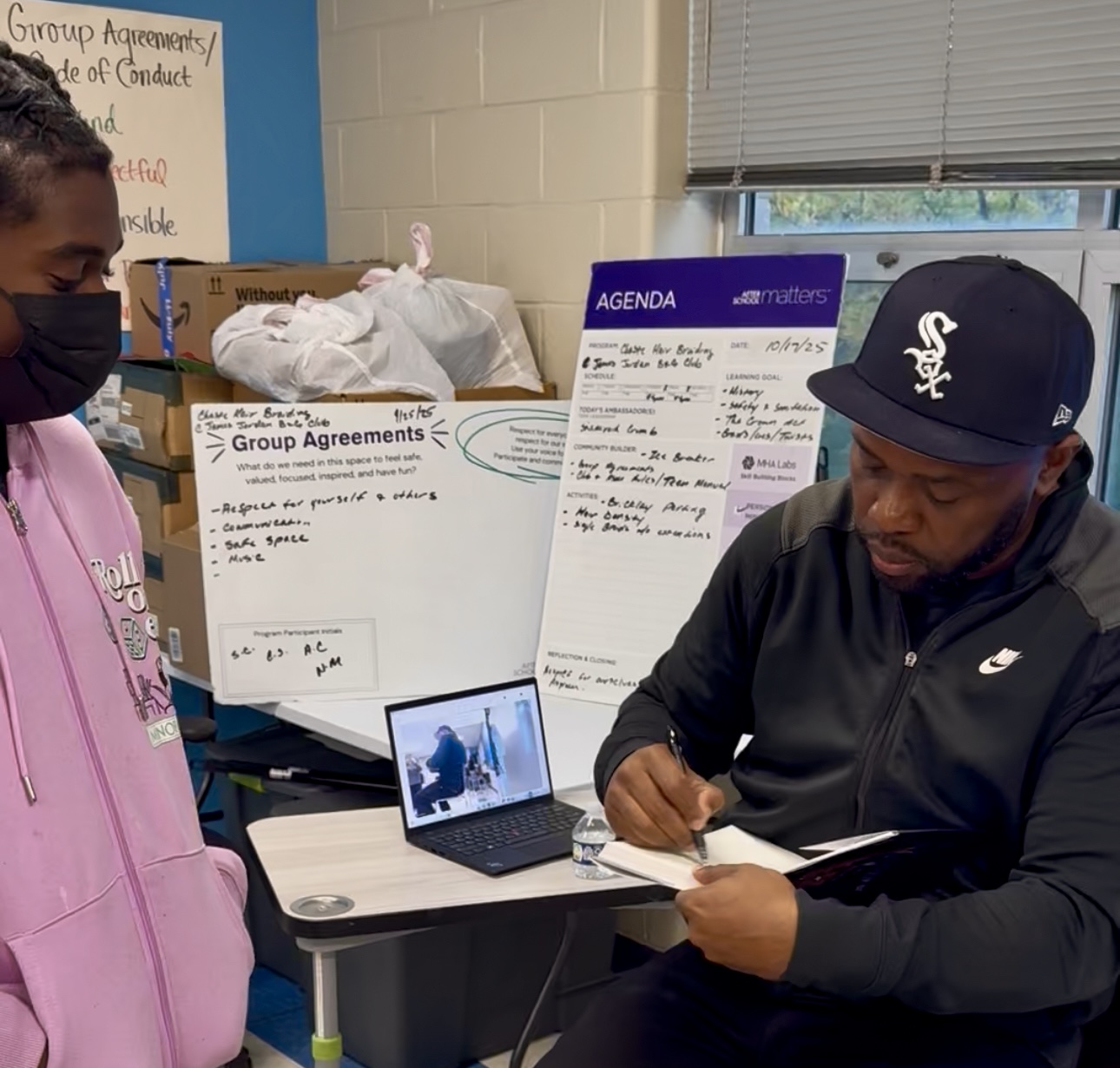 A man in a black tracksuit and cap signs a book for a girl in a pink hoodie. Behind them are whiteboards with notes, a laptop, and boxes.