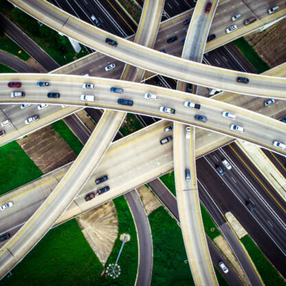 Aerial view of a busy multilayered highway interchange with cars traveling in multiple directions, surrounded by patches of green grass and brown fields.