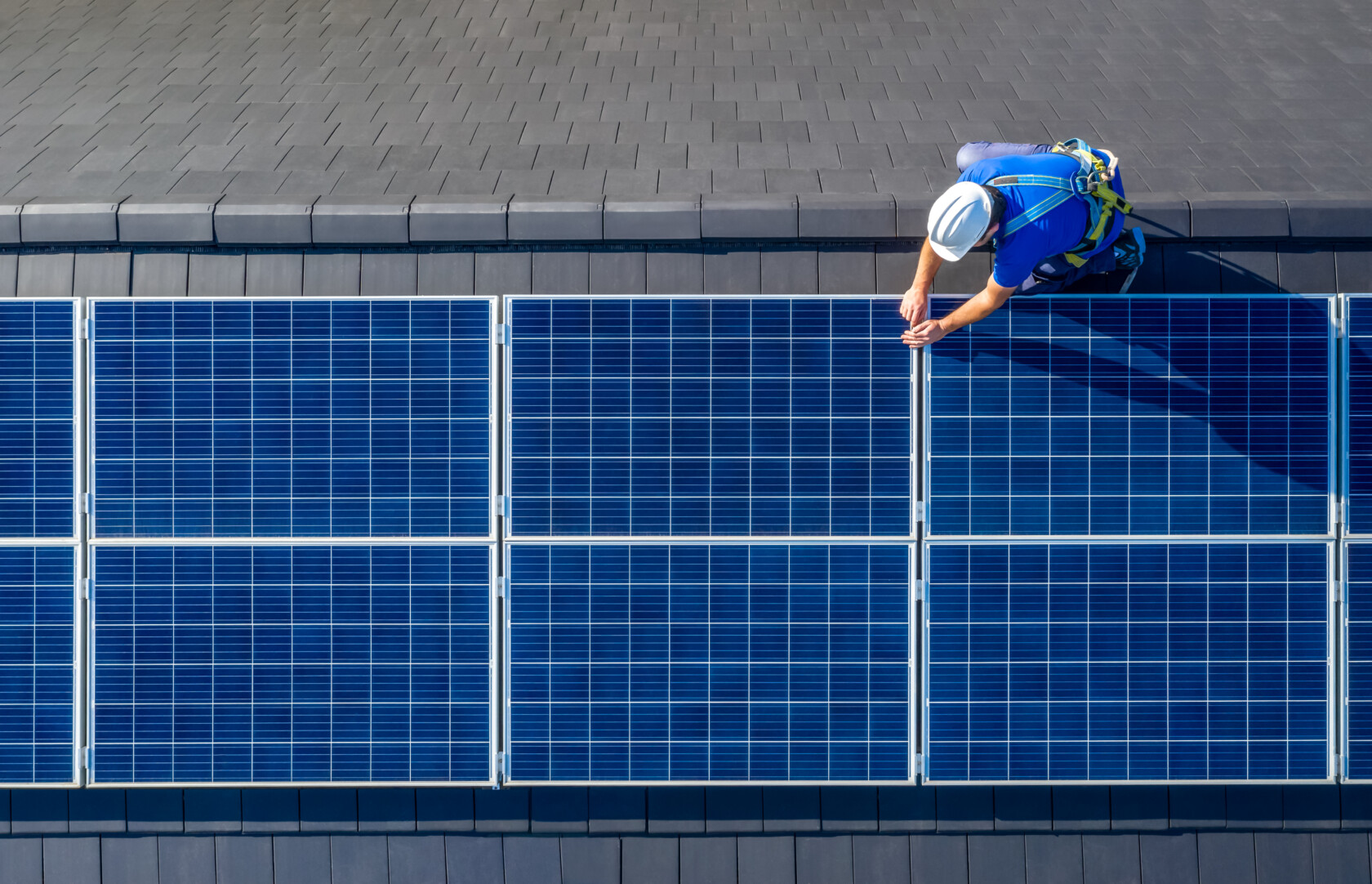 A worker in safety gear installs or inspects blue solar panels on a sloped gray tiled roof.